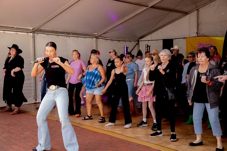 Maddison Glover leading a mixed-age crowd through a line dance under a marquee, microphone in hand.