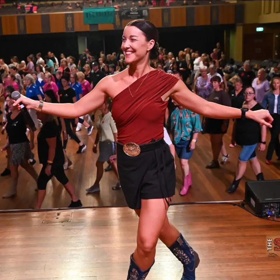 Maddison Glover teaching a line dance, microphone in hand, cowboy boots on a wooden floor in front of a hall of dancers.